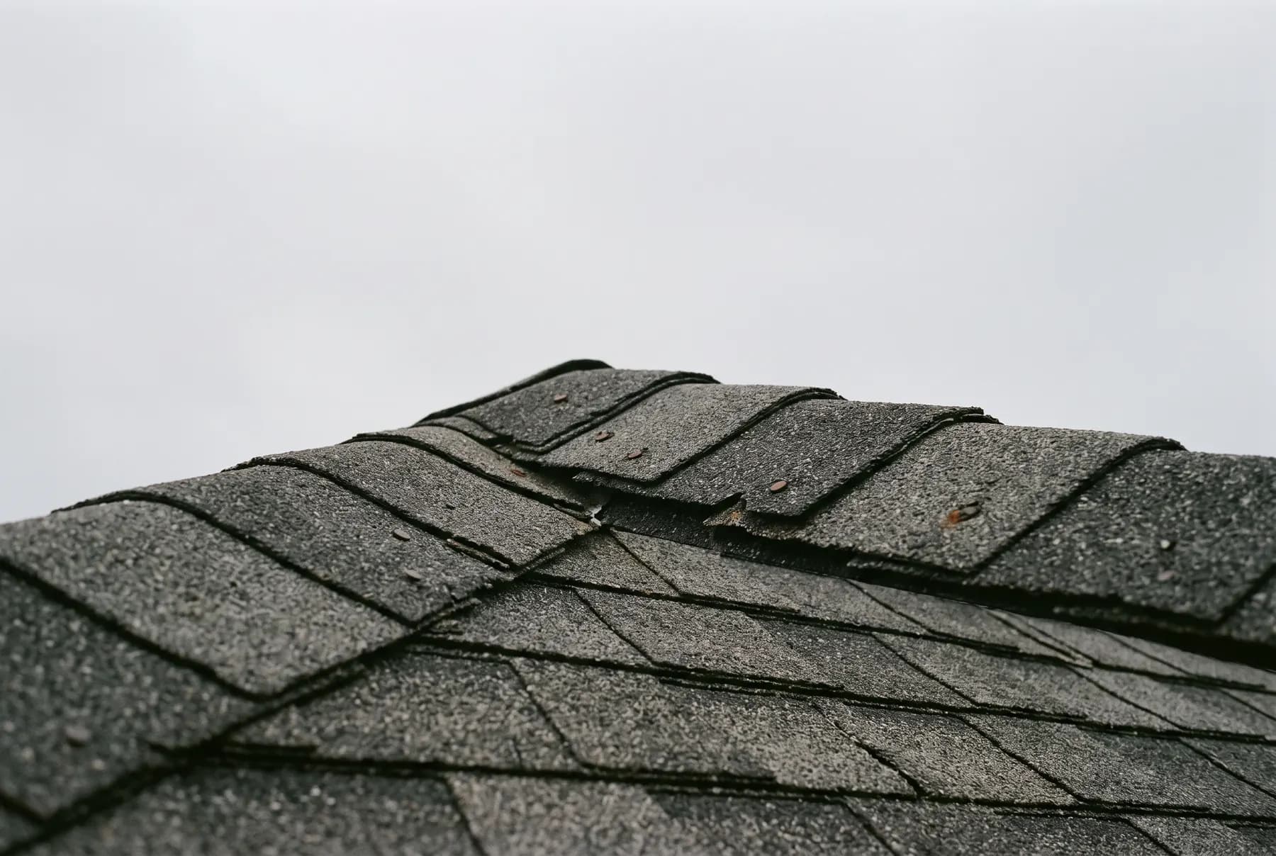 A roof ridge cap detail on architectural shingles against an overcast sky.